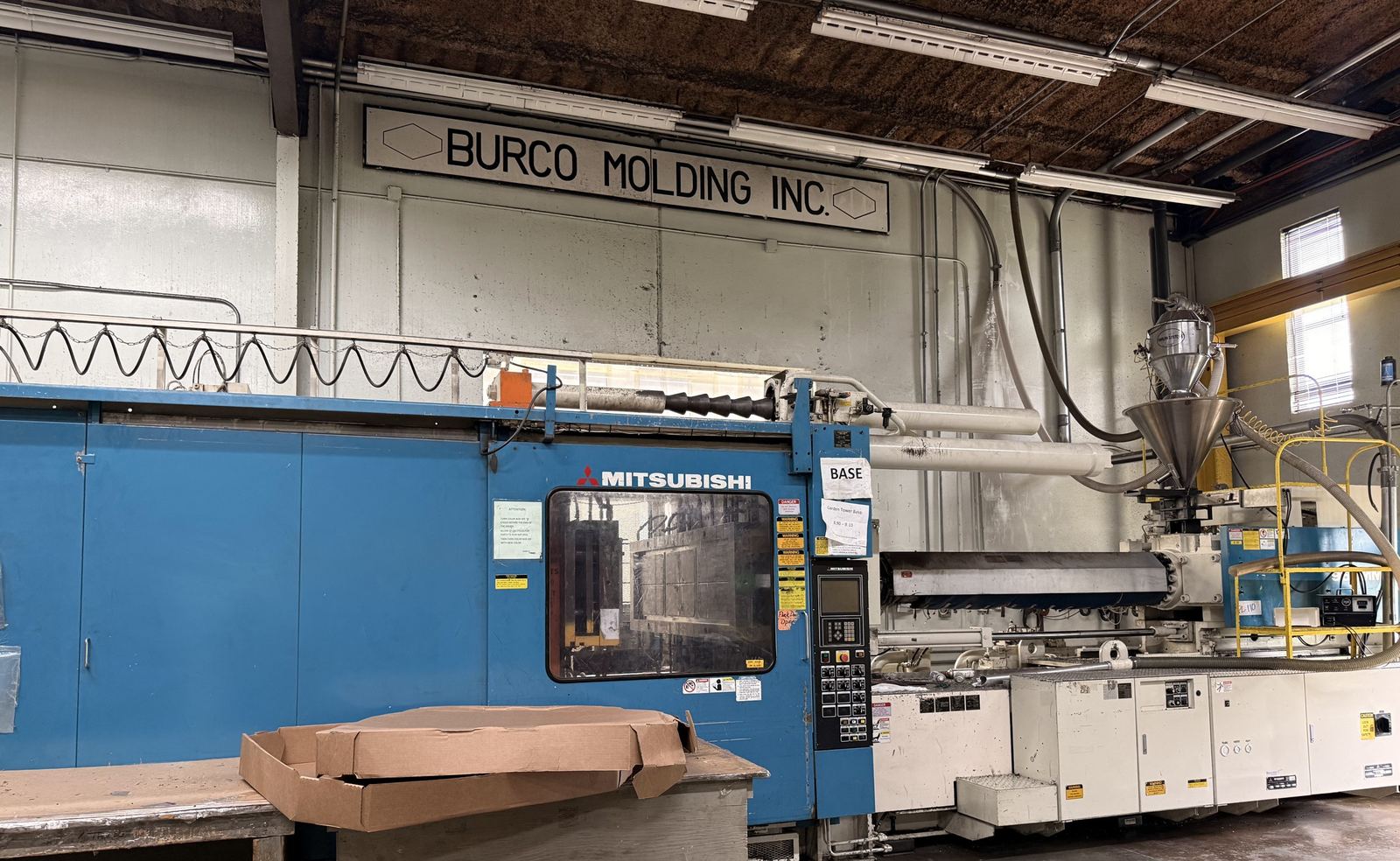 Blue Mitsubishi injection molding machine under the painted Burco Molding Inc. wall sign, hopper and dryer at right