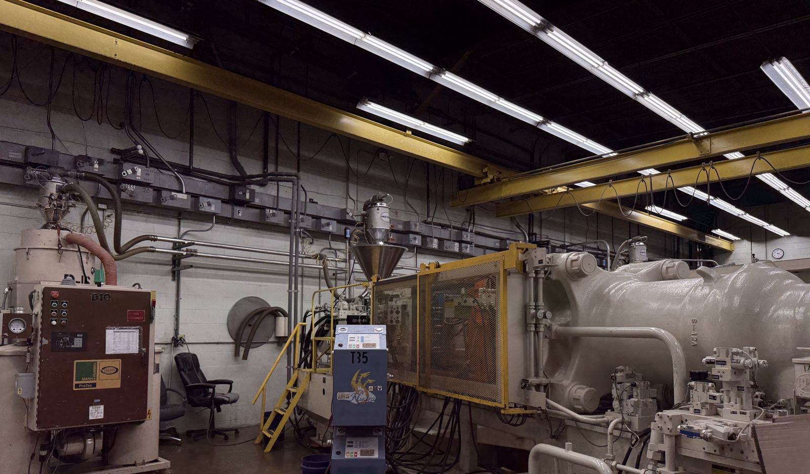 A large white injection molding machine with a yellow guard door, dryer and loader beside it, under the bridge crane