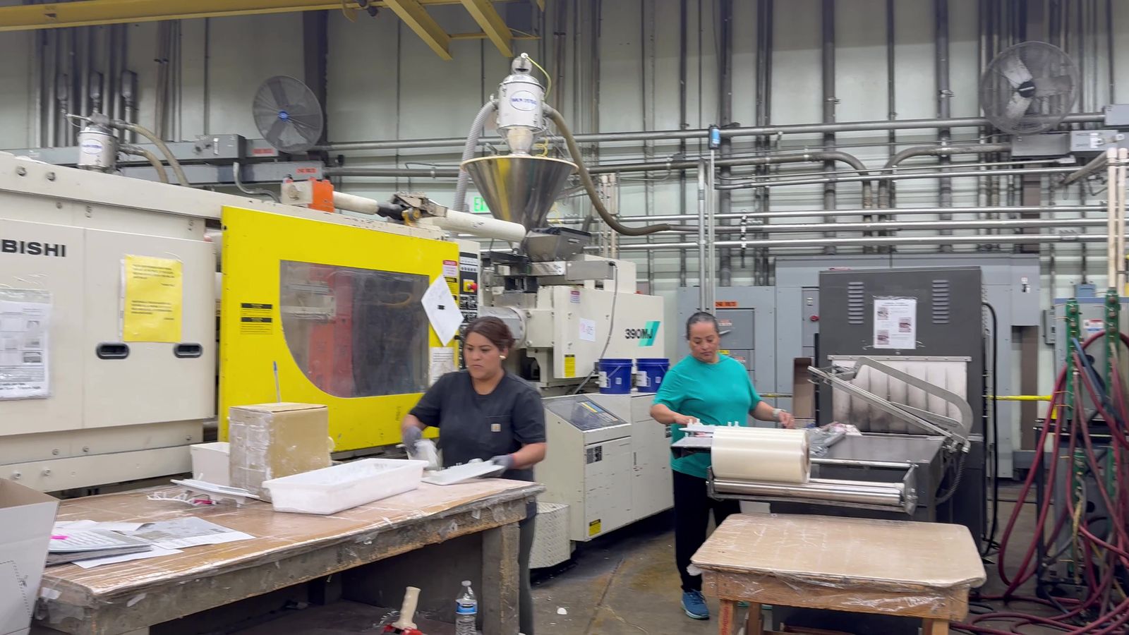 Two Burco operators at a bagging line beside a yellow injection molding machine, one trimming parts and one sealing bags