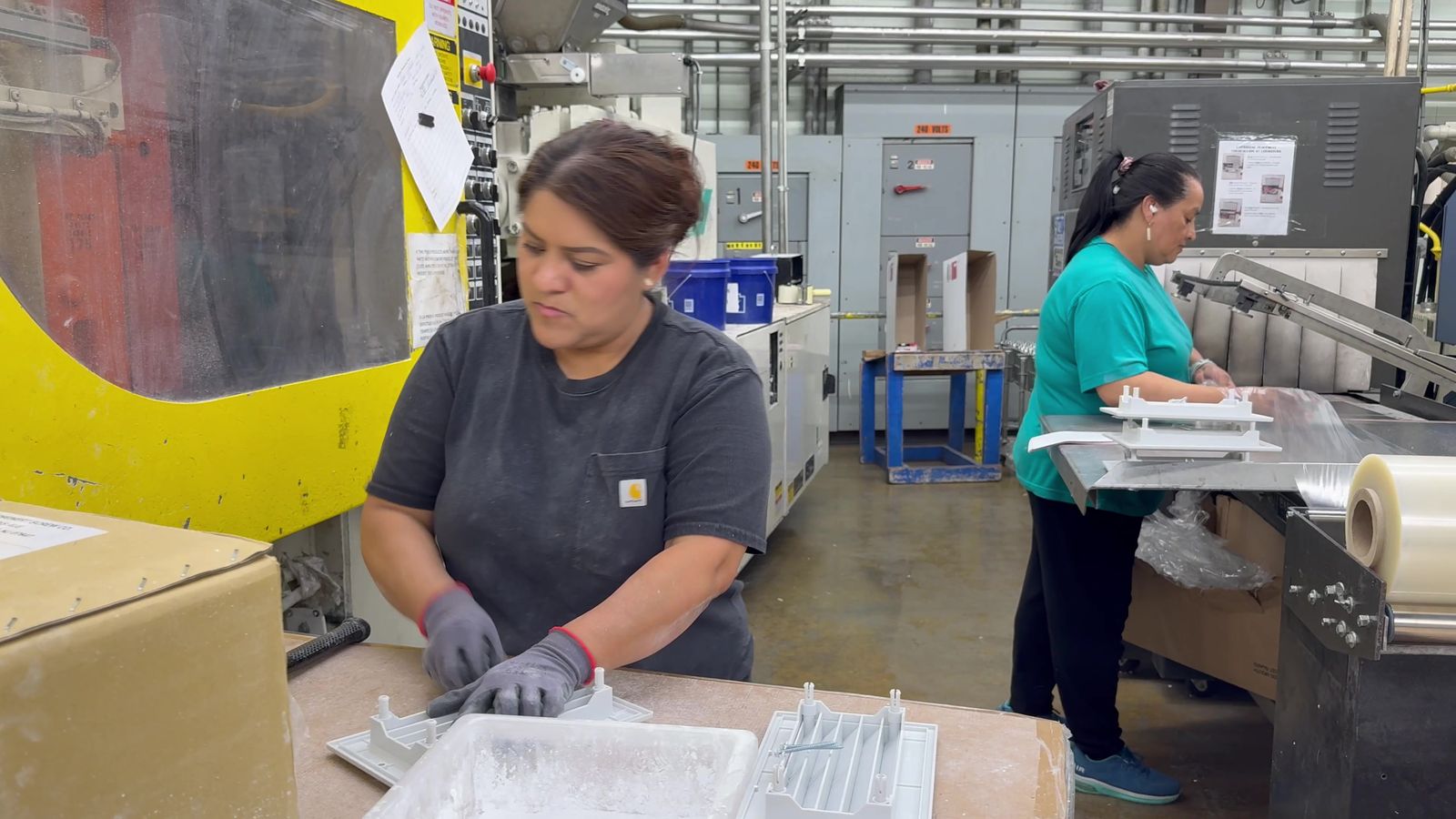 A Burco operator in work gloves trimming and inspecting white plastic parts at a packing table beside a yellow injection molding machine