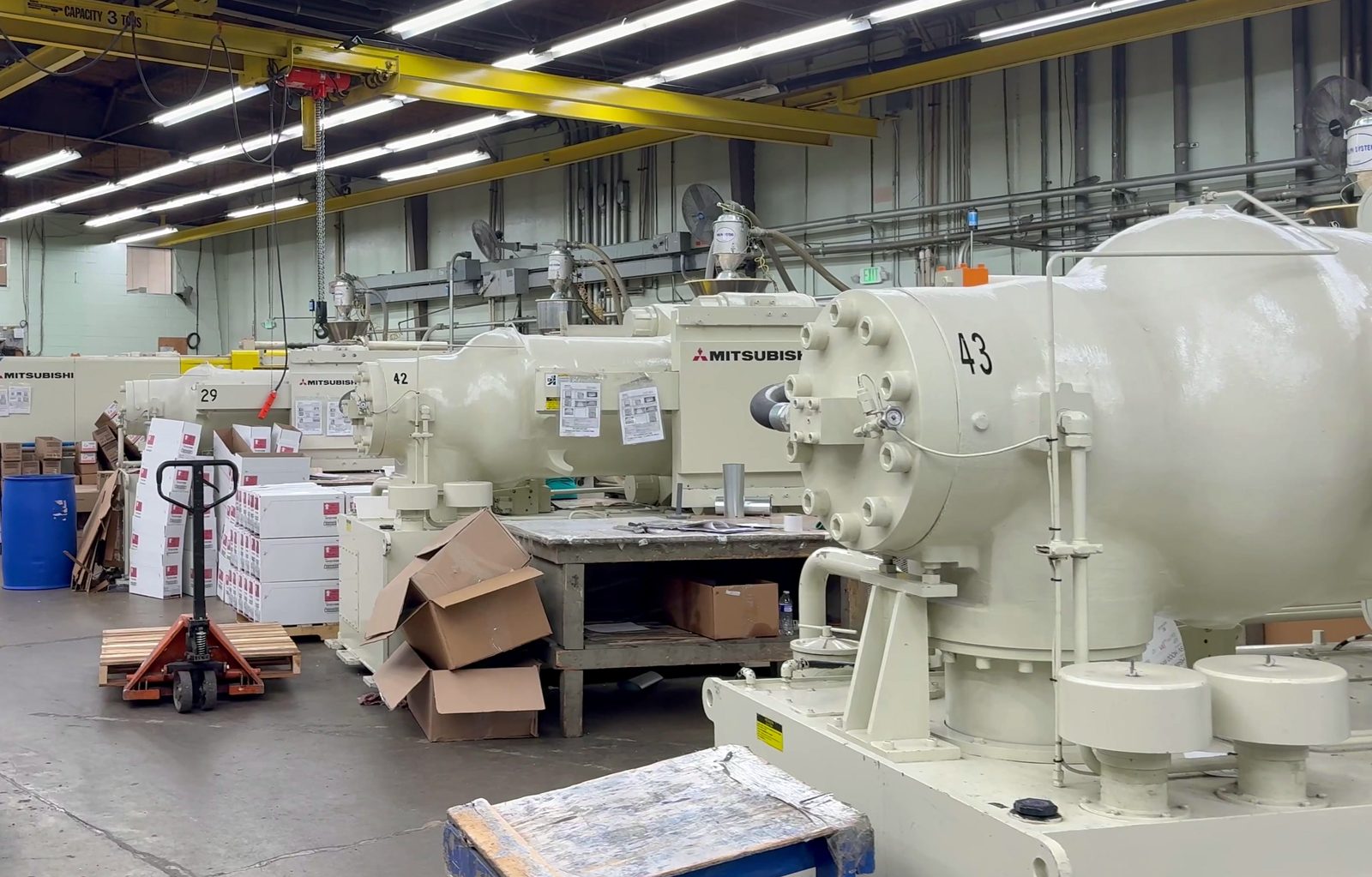 Large white Mitsubishi machines in a row on the Burco floor, machine 43 in front, a yellow bridge crane overhead